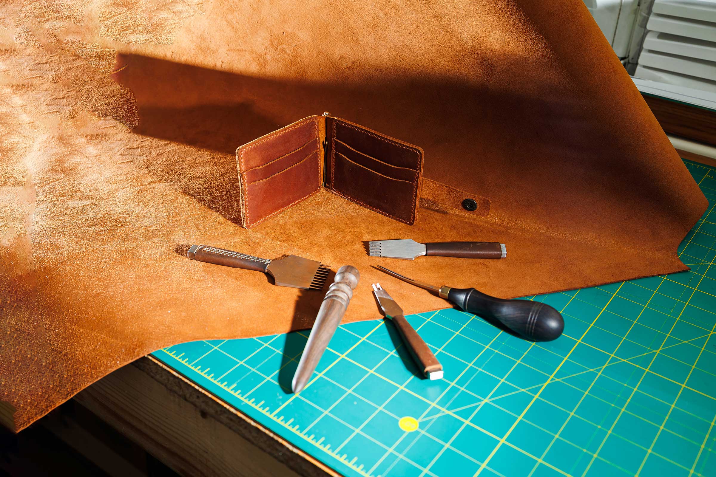 Raw leather on cutting table with leather tools and a finished leather wallet.