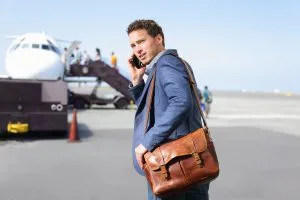 Man on airport runway on phone carrying leather shoulder bag about to board airplane.