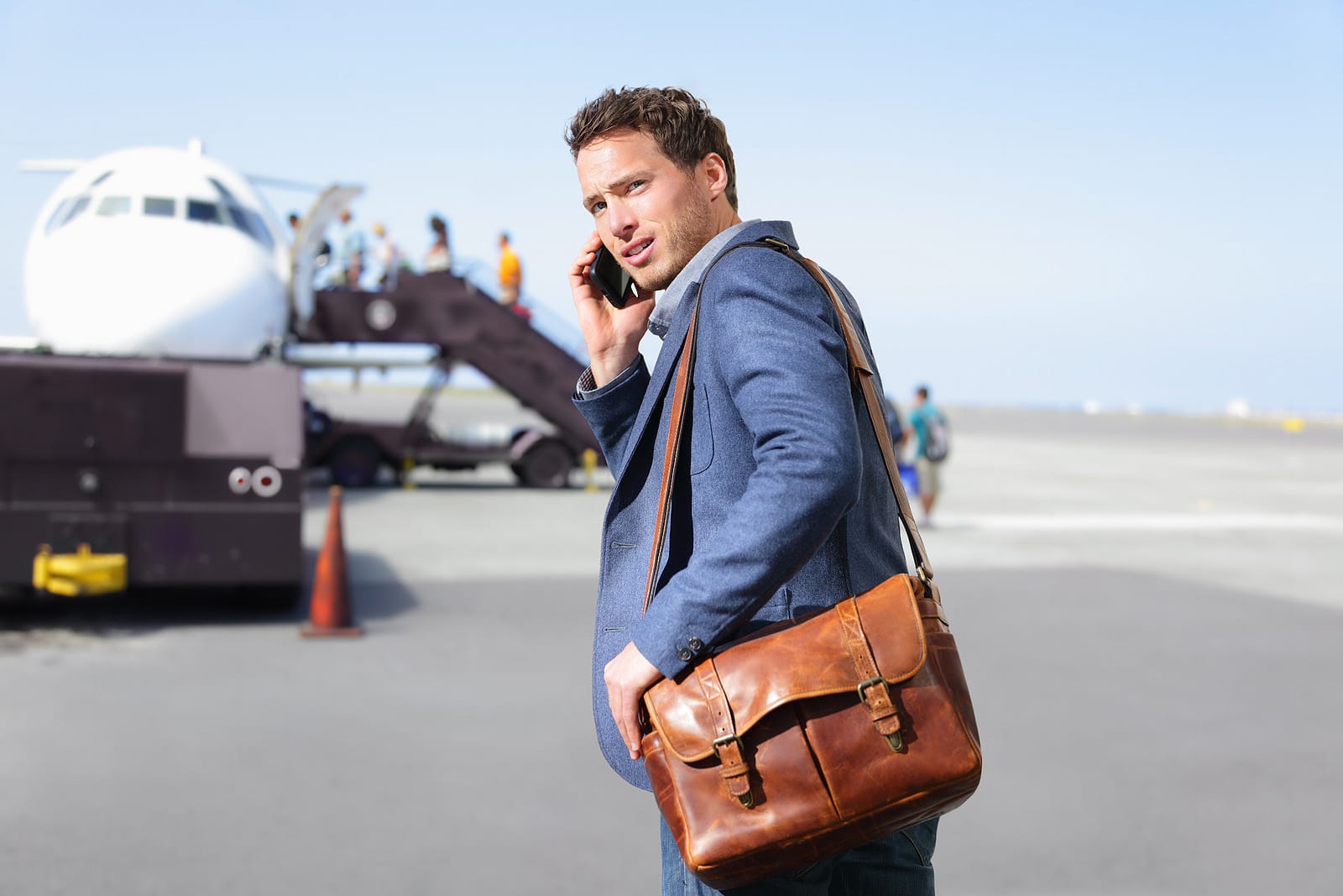 Man on airport runway on phone carrying leather shoulder bag about to board airplane.