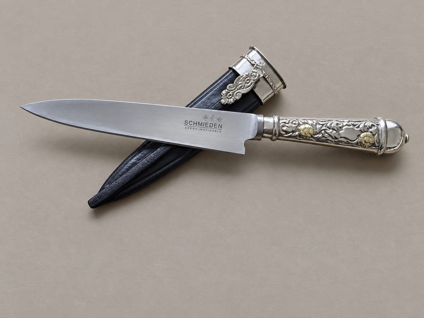 Argentine gaucho knife with an engraved alpaca handle and small gold-toned floral accents, displayed over its black leather sheath