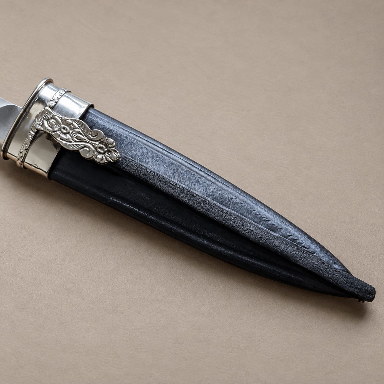 Close-up of a black leather Argentine knife sheath with a silver-toned alpaca fitting and floral tab