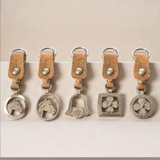 Five handcrafted Argentine key rings displayed side by side, each featuring a natural leather loop and polished Alpaca silver motif. The collection includes two horse head designs, a Criollo stirrup, a square floral medallion and a round floral medallion.